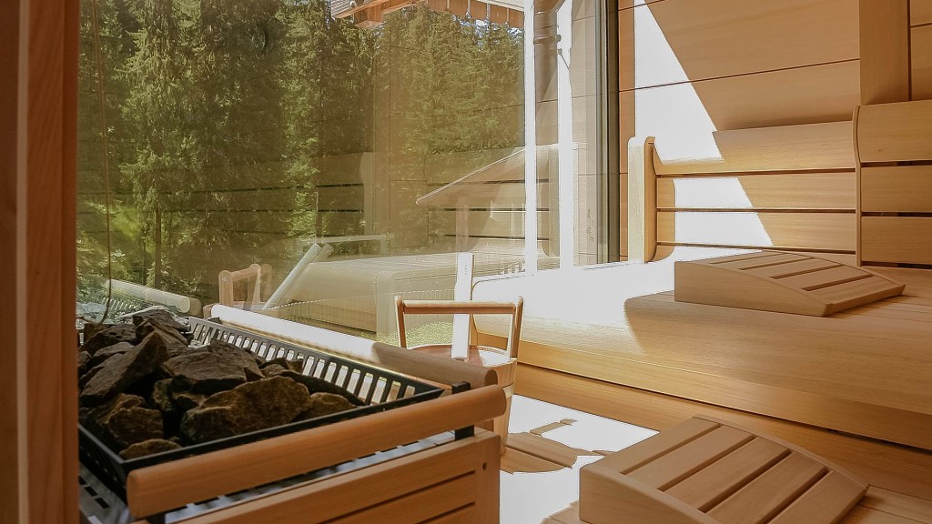 Interior of a Finnish sauna featuring wooden benches and a stone heater, with large windows offering a view of lush green trees outside.
